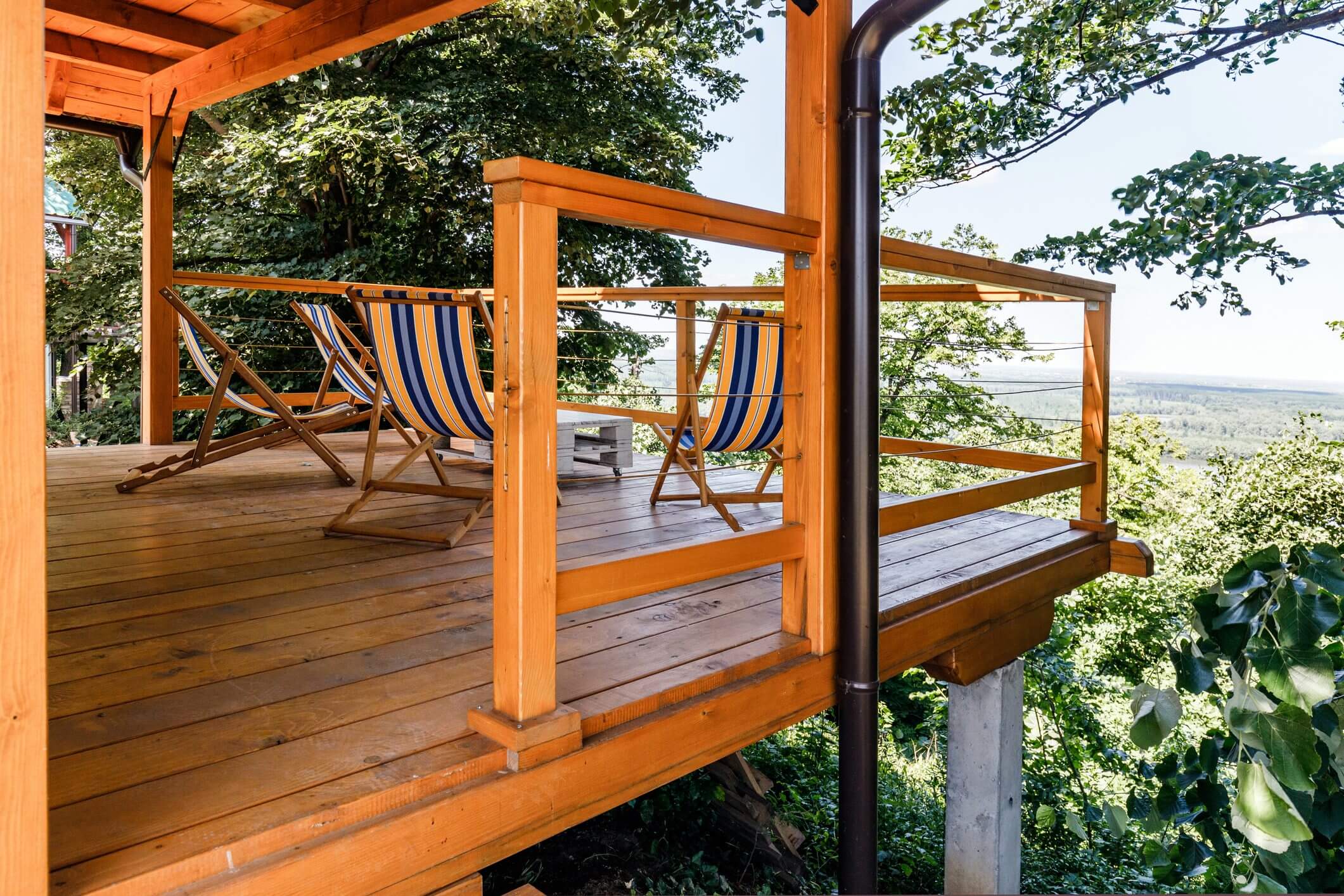 Wooden deck with glass railing and colorful lounge chairs overlooking greenery.
